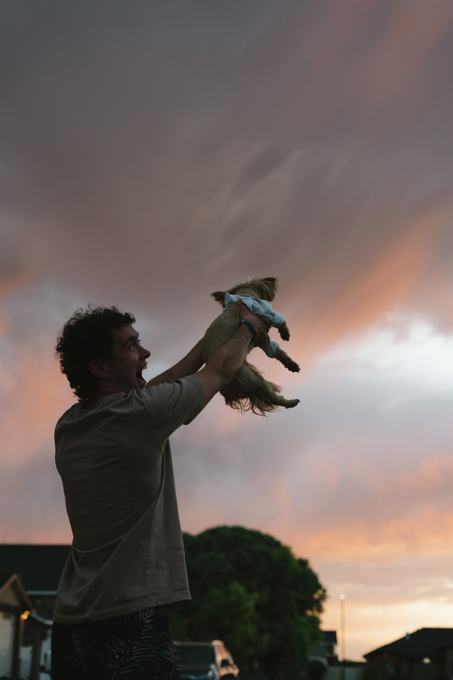 Man lifting a small dog in a shirt toward a pink and orange sunset sky