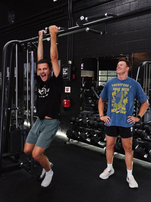 Two friends at a dark gym, one hanging from a pull-up bar in a Made New shirt