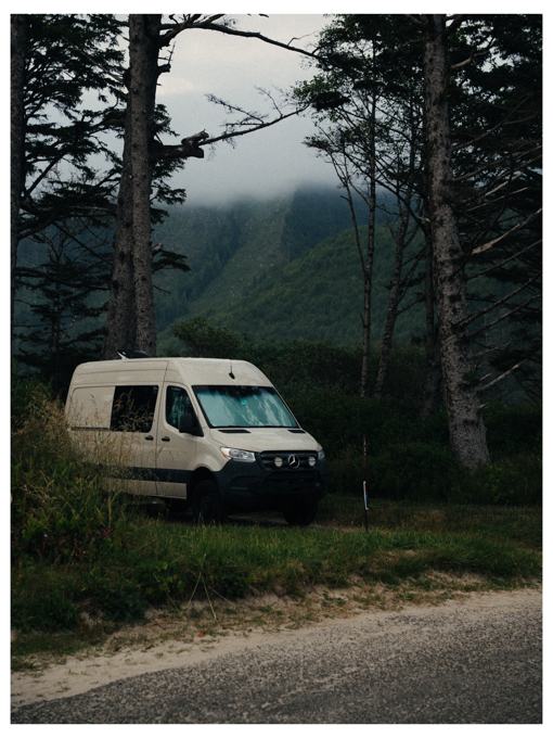 Camper van parked under tall trees with misty mountains behind