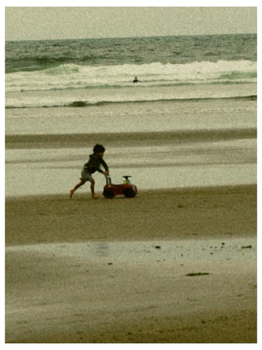 Child pushing a toy car along a grainy beach with a surfer in the waves