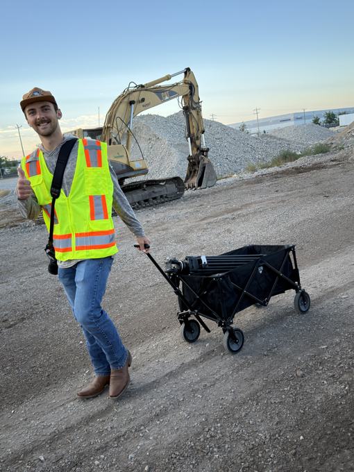 Trace Keller in a safety vest pulling a camera gear wagon past an excavator