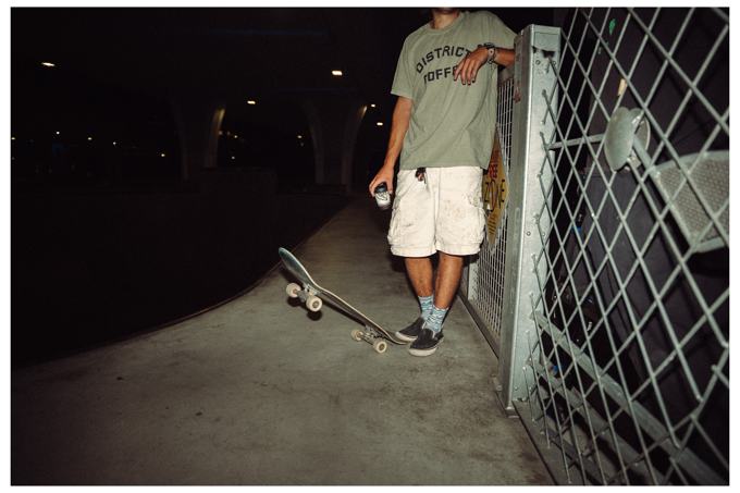 Skater leaning on a fence at night with a coffee, board tipped on the ground