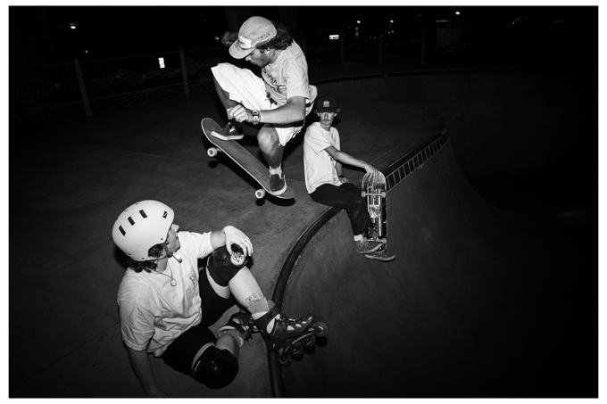 Black and white skate bowl at night, a skater mid-air over a rollerblader and a friend sitting on the edge