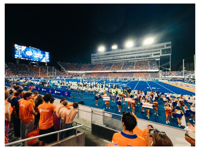 Boise State football stadium on the blue turf at night, fans in orange and cheerleaders on the sideline