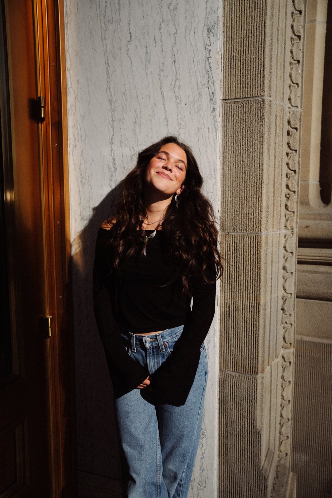 Adeline with long dark curls leaning against a marble wall in warm sunlight, eyes closed and smiling, wearing a black top and jeans