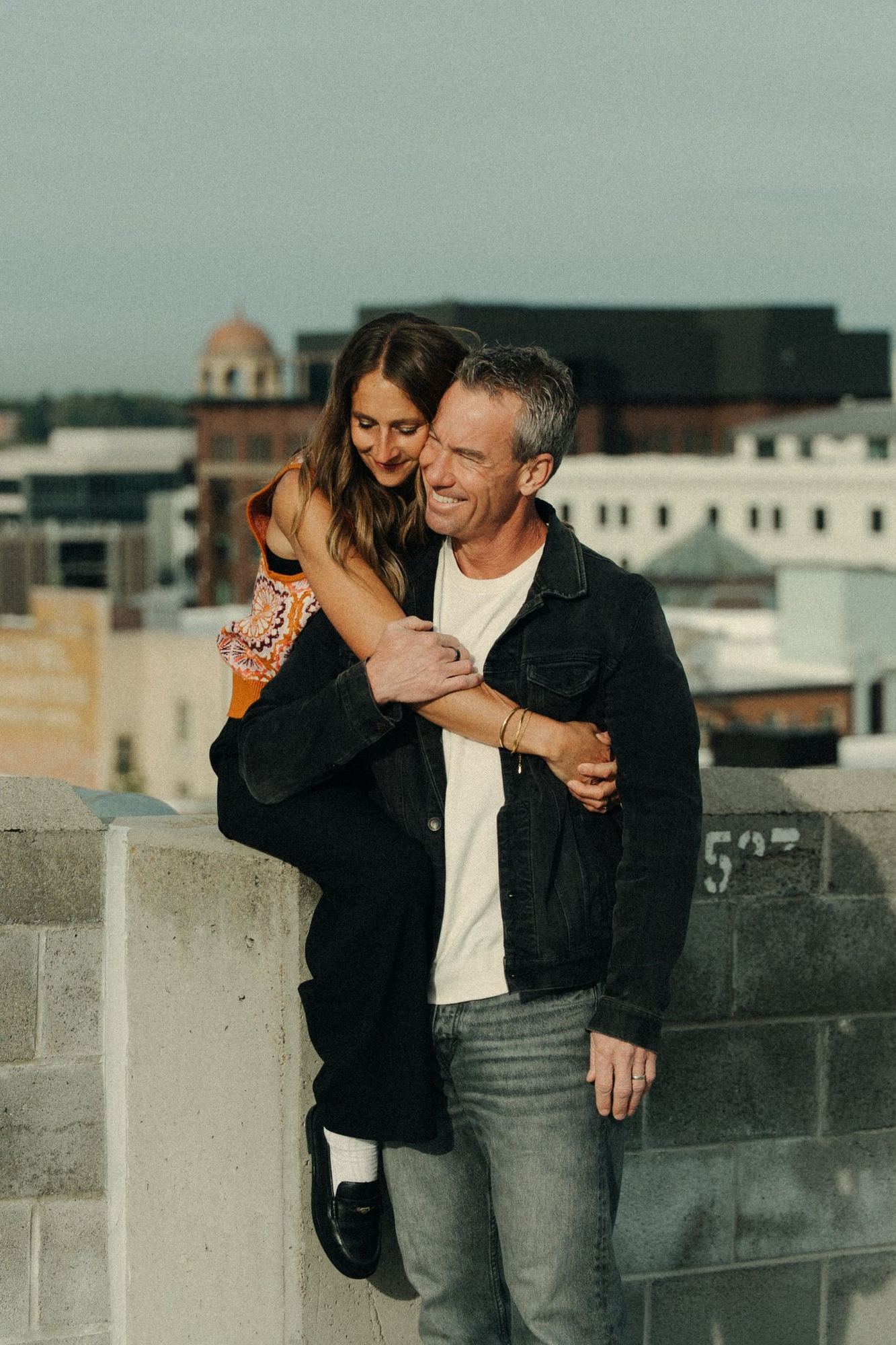 Couple embracing on a Boise rooftop at golden hour, her arms around his shoulders, with downtown Boise buildings behind them