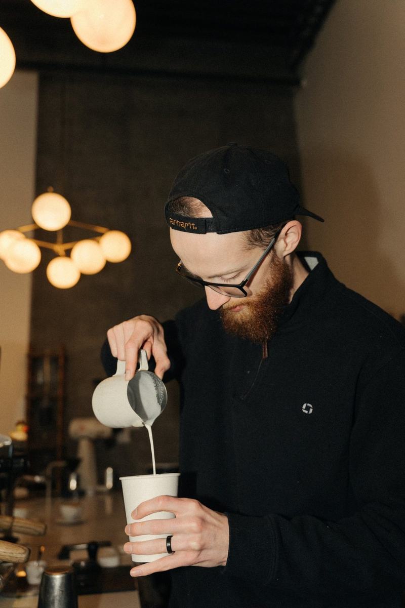A barista pouring steamed milk into a paper cup at a coffee bar with warm globe lights behind him
