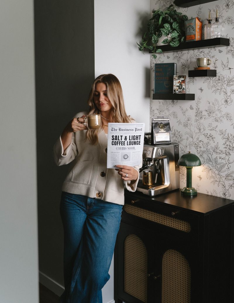 A woman in a cream cardigan sipping a latte and holding a Salt & Light Coming Soon newspaper beside an espresso machine, green lamp and floral wallpaper