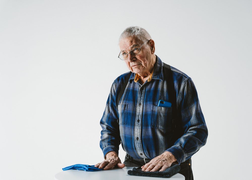 An older man with gray hair and glasses in a blue plaid flannel shirt and suspenders resting his hands on a white table with two folded towels, against a clean white studio backdrop