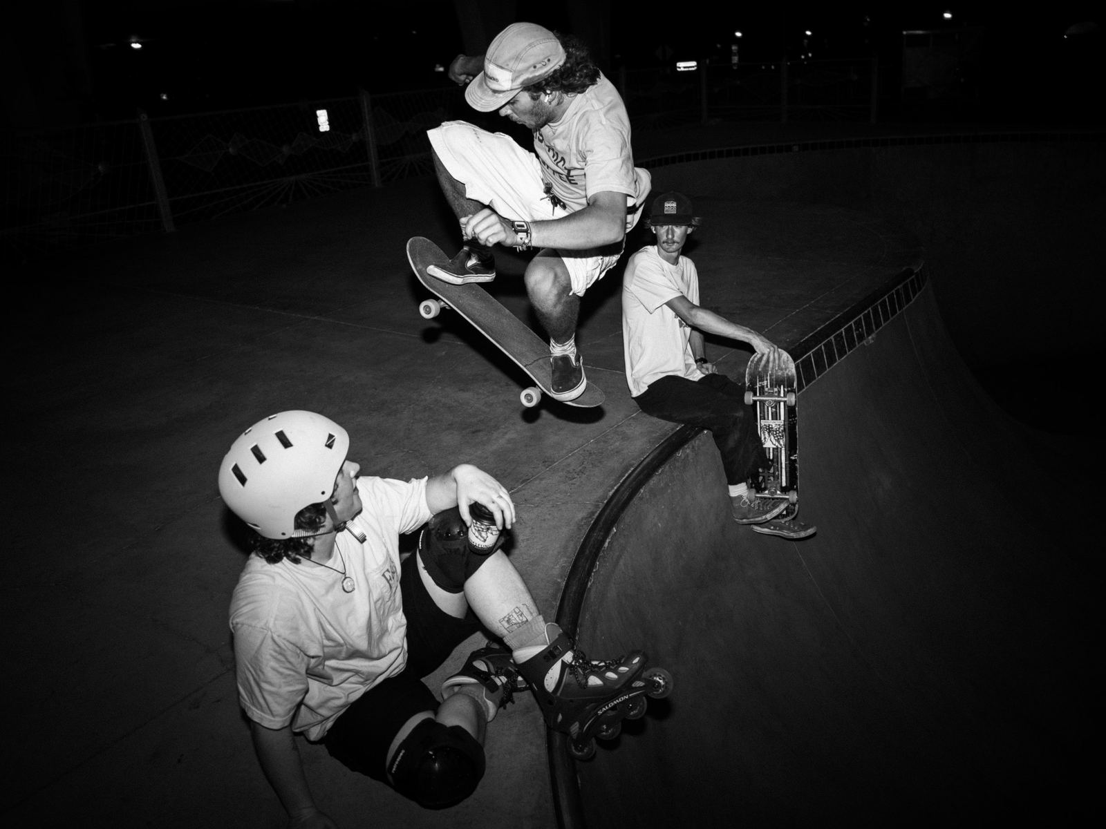 Black and white photo of a skateboarder jumping across a skate bowl at night while two friends watch from the edge
