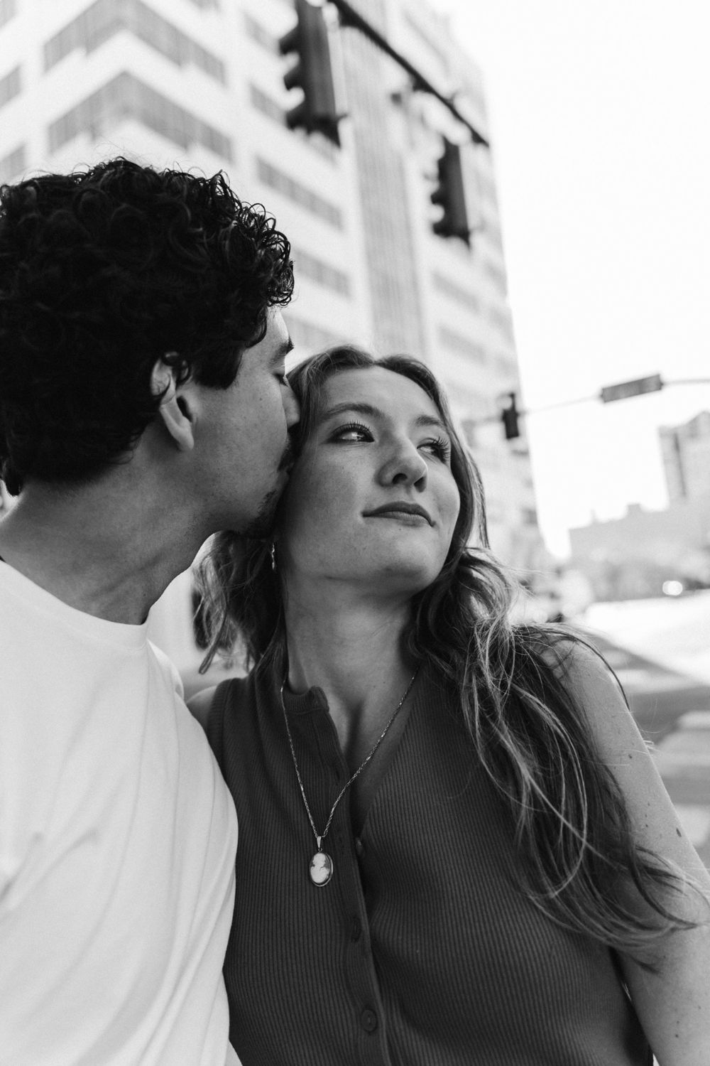 A man in a white t-shirt kisses his partner on the forehead as she glances off to the side, downtown Boise buildings behind them, black and white