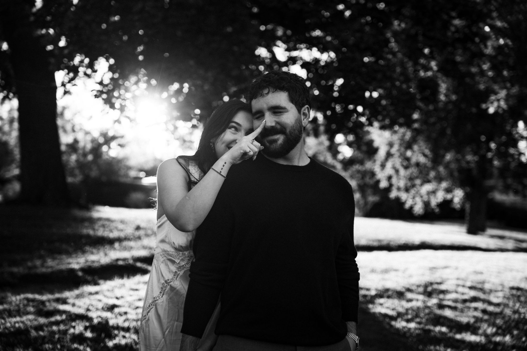 Black and white photo of Cora playfully tapping James on the nose in backlit sunlight under the trees