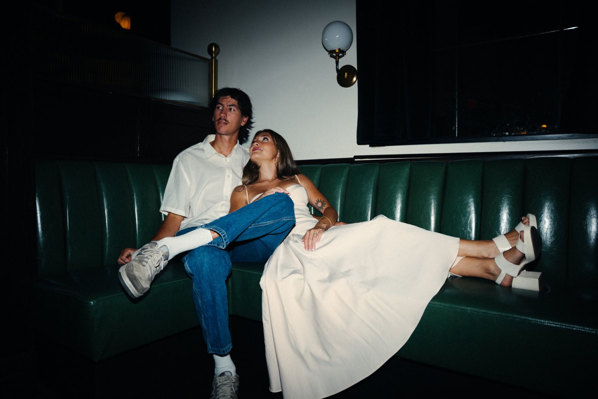 Audry leaning back against Lucas on a curved green leather booth, her legs stretched out in white platform sandals