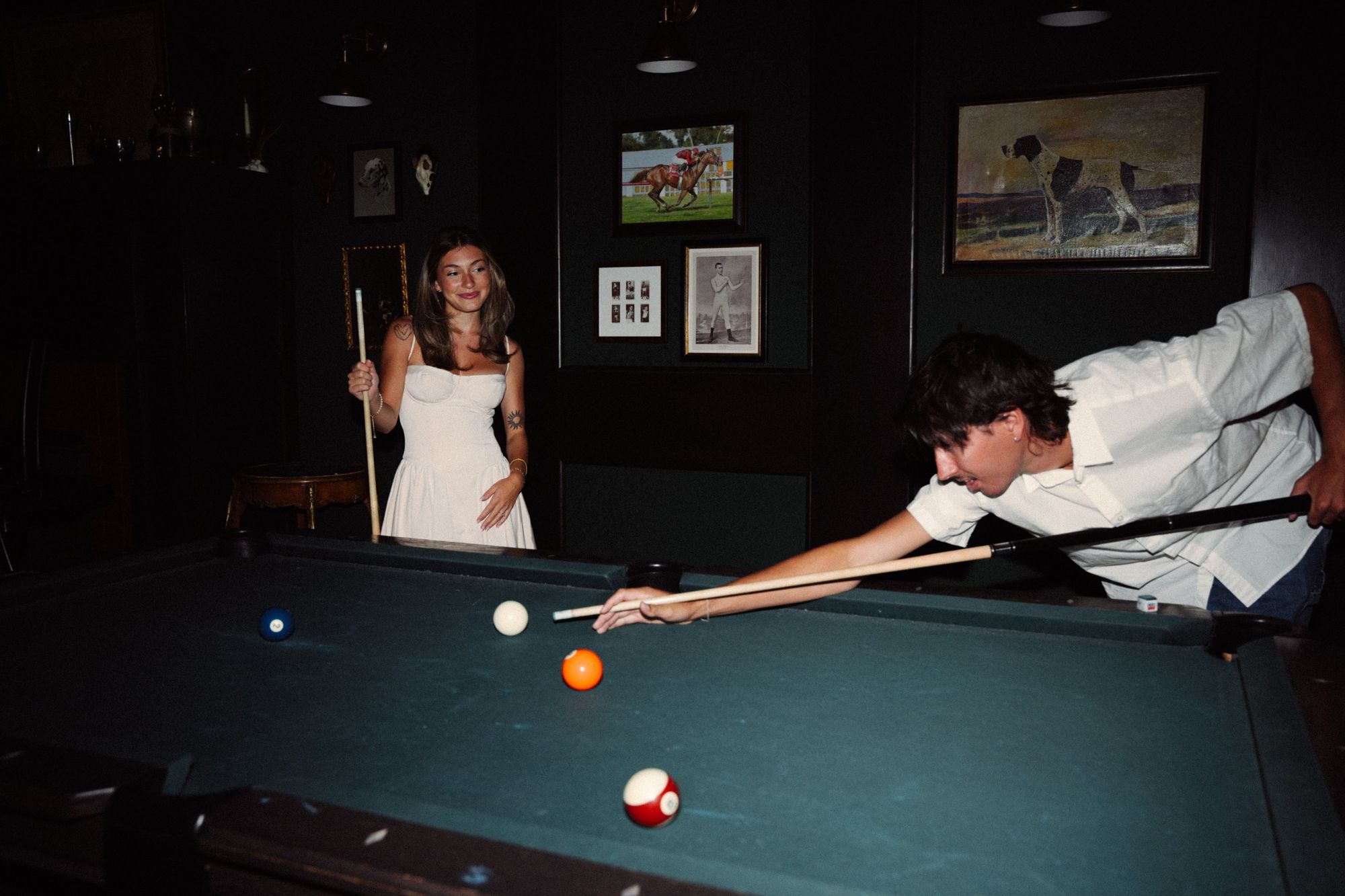 Lucas lining up a shot at a pool table while Audry watches and smiles, in a dark green room with framed art