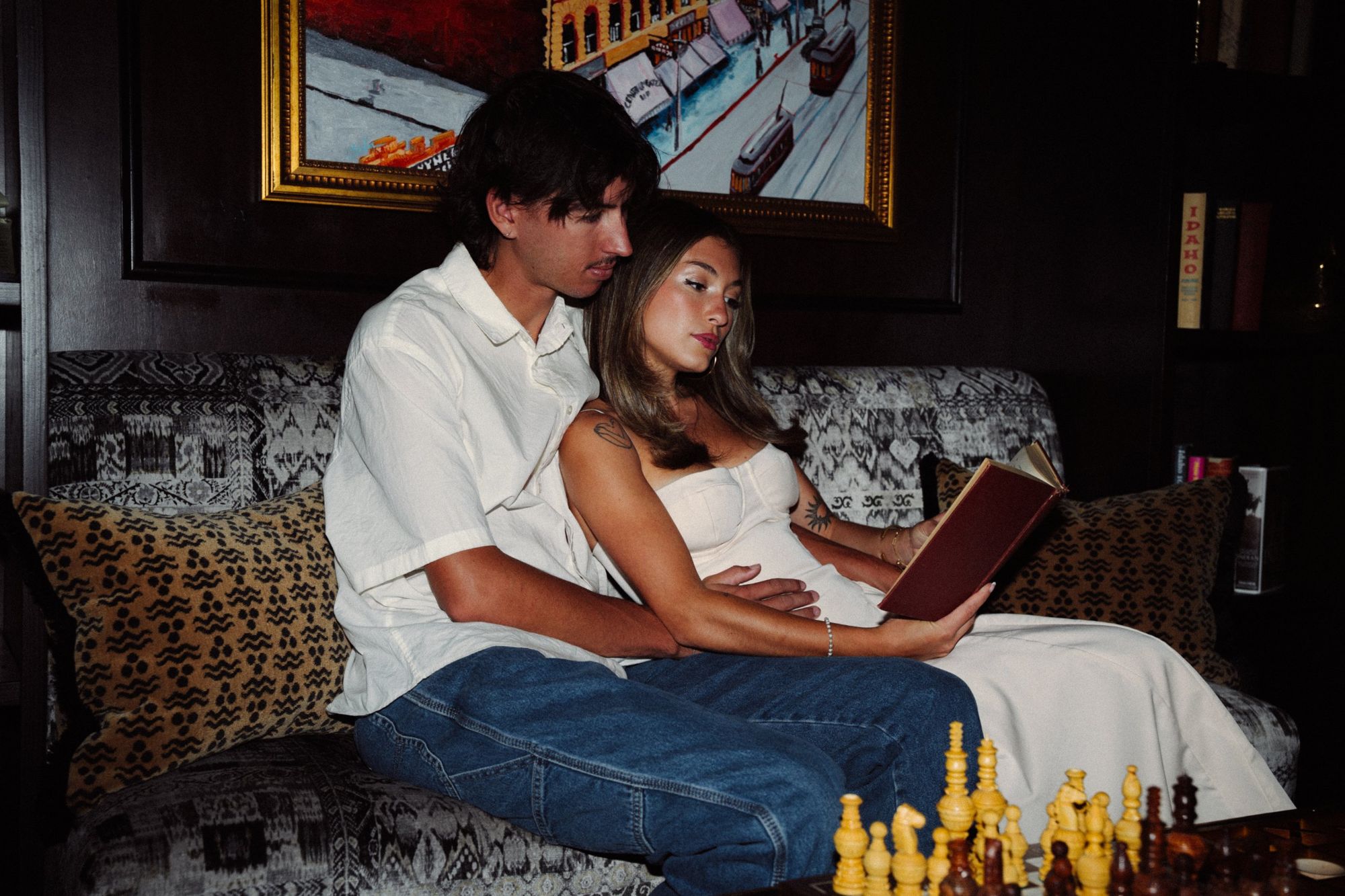 Audry holding an open red book while Lucas hugs her from behind on a patterned sofa, a chess board in the foreground