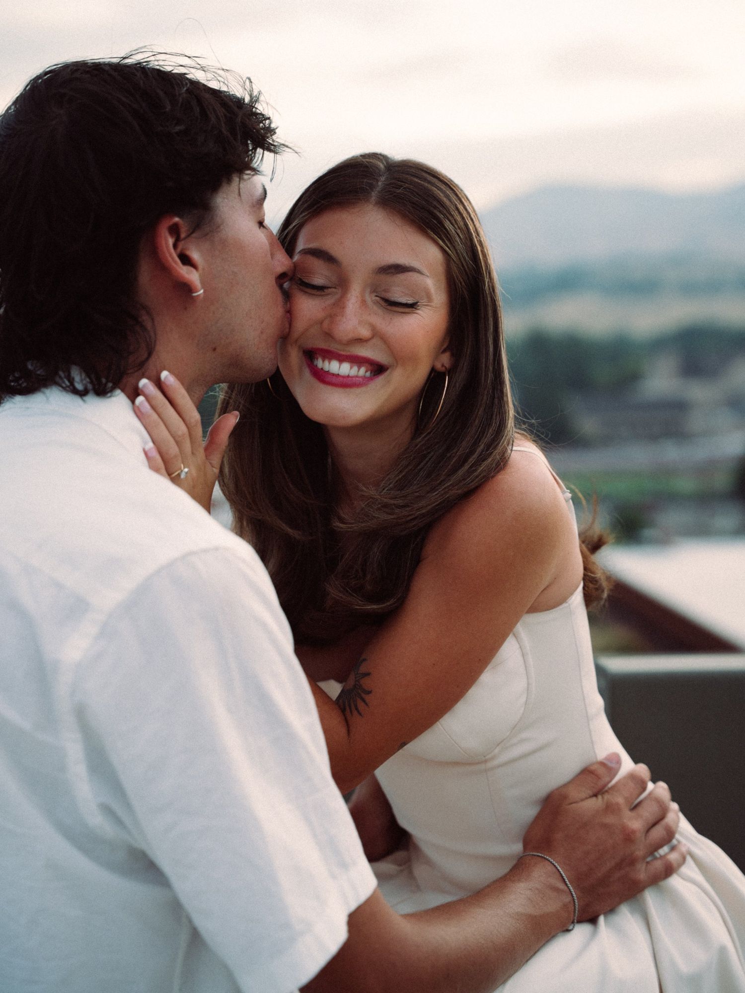 Lucas kisses Audry's cheek as she laughs with her eyes closed, her hand on his neck, on a rooftop at dusk