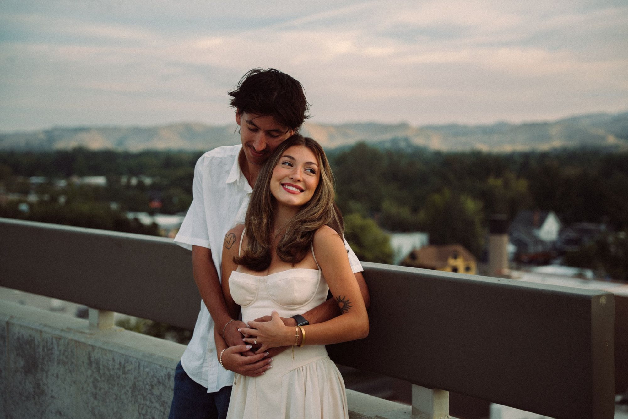 Lucas hugs Audry from behind on a rooftop while she smiles off to the side, with the Boise foothills behind them