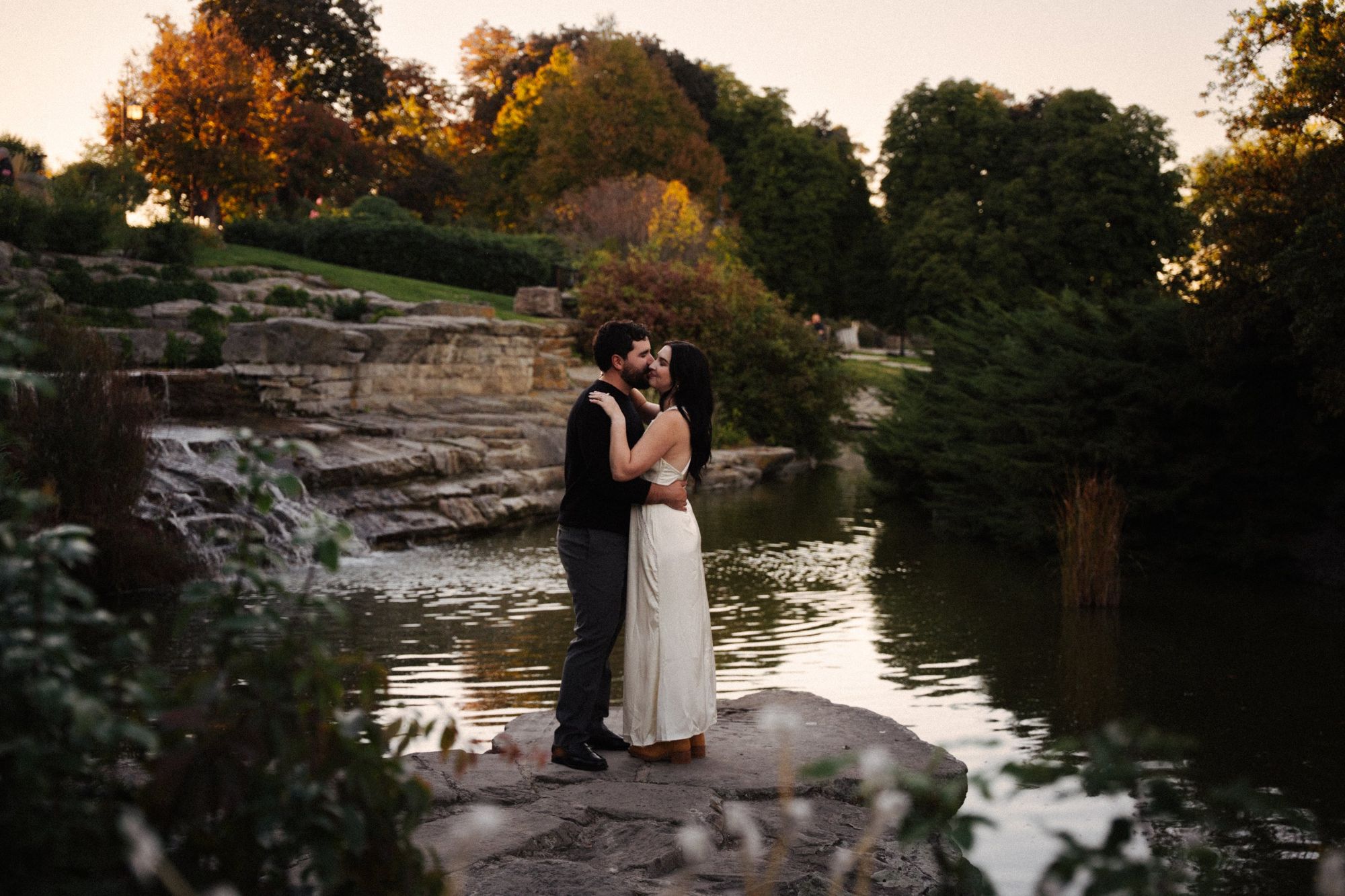 James and Cora kiss on a flat rock beside a pond and small waterfall, surrounded by fall foliage
