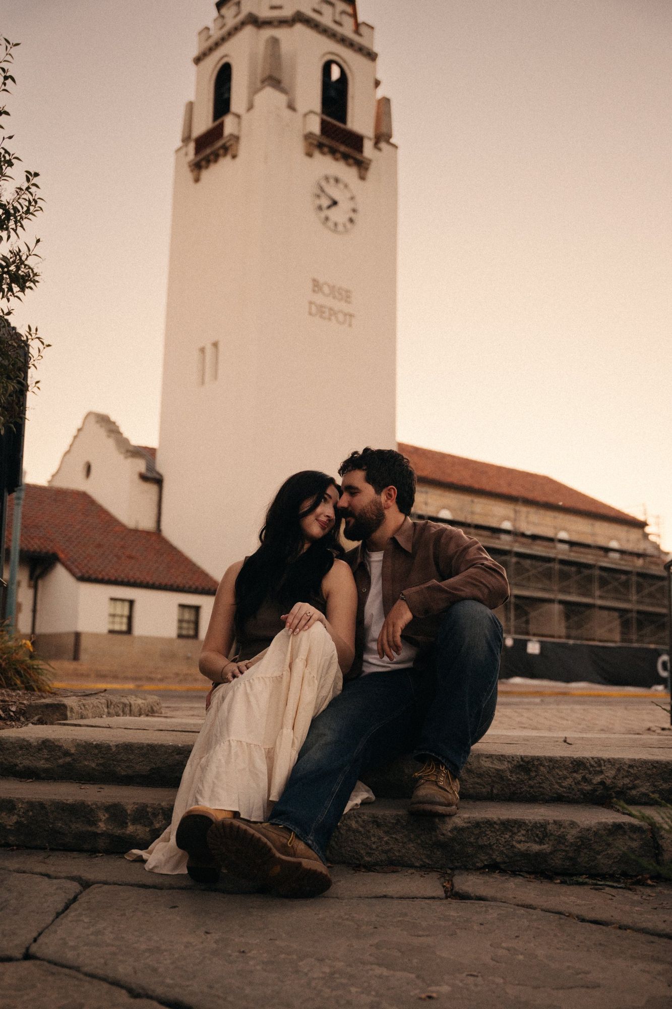 James and Cora sitting close on stone steps in front of the Boise Depot clock tower at sunset