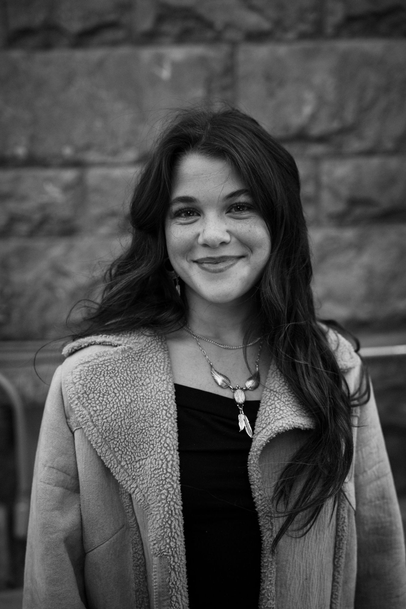 Adeline smiling at the camera in black and white, wearing a shearling coat and feather necklace in front of a stone wall