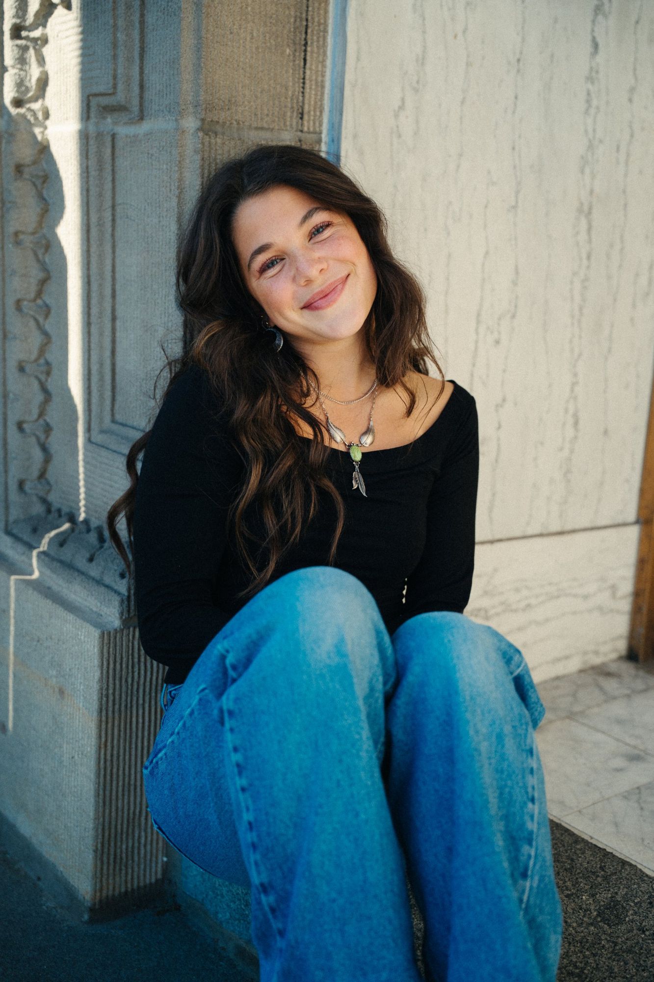 Adeline sitting against a stone wall with her head tilted, smiling, in a black top, jeans and a feather necklace