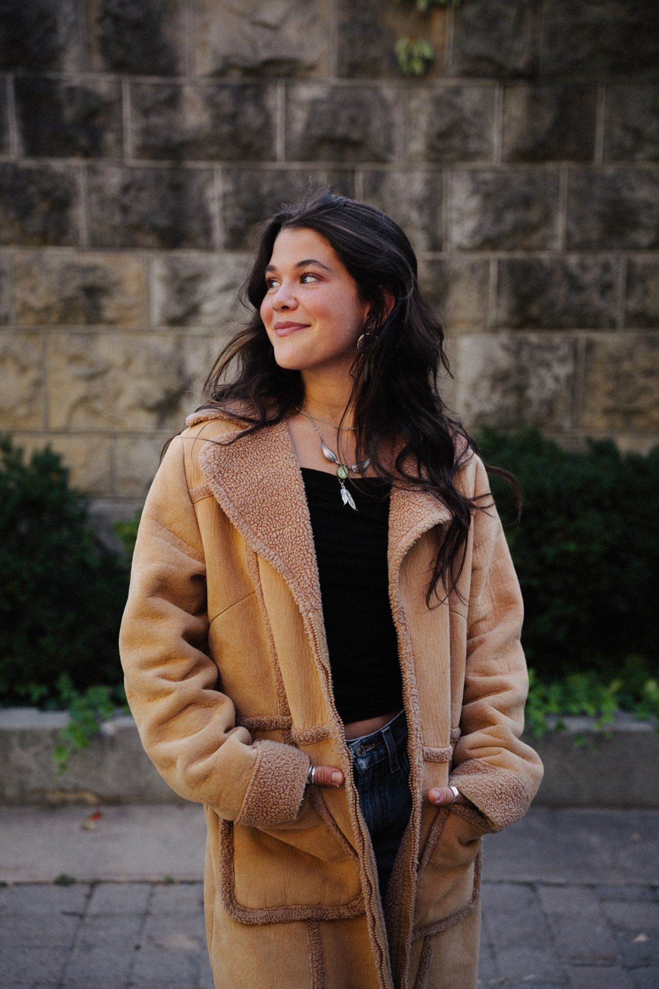 Adeline looking off to the side with a smile, hands in the pockets of a tan shearling coat, in front of a stone wall