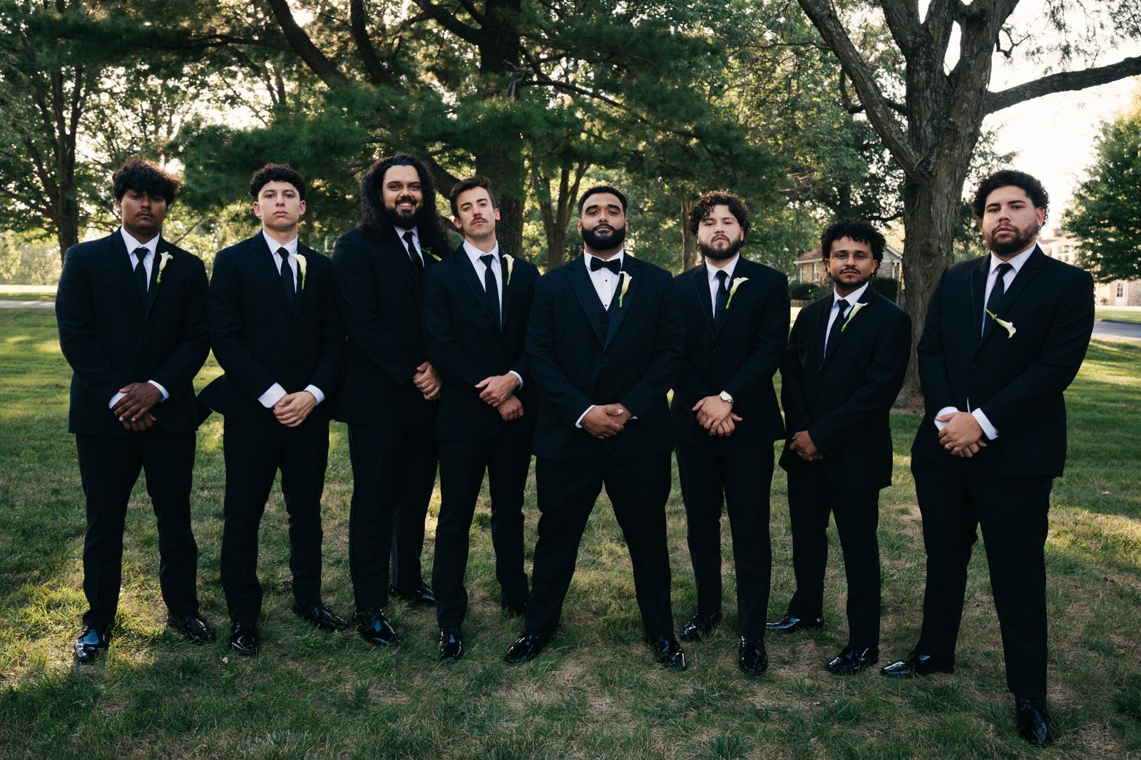 Caio and his eight groomsmen in black suits with white calla lily boutonnieres standing in a row under tall trees