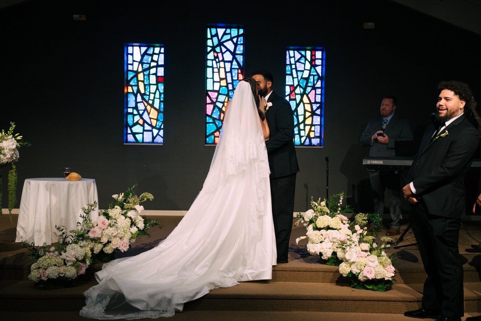 Caio and Trinity kiss at the altar in front of three stained glass windows, her long lace veil trailing across the stage