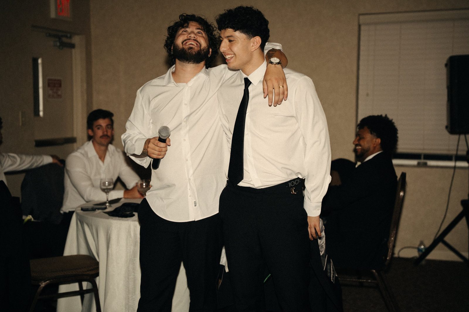 Two guests laughing with their arms around each other, one holding a microphone, at the wedding reception