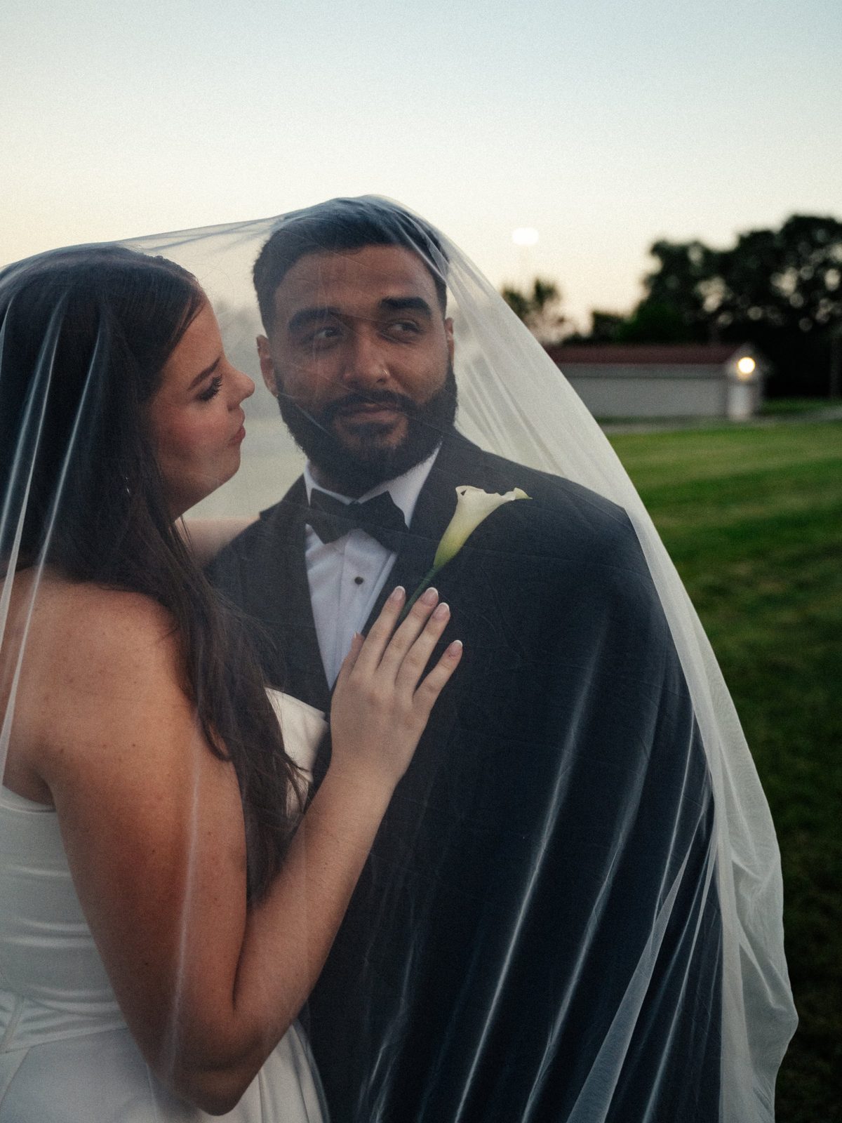 Trinity leaning in close to Caio while they stand together under her sheer veil at dusk