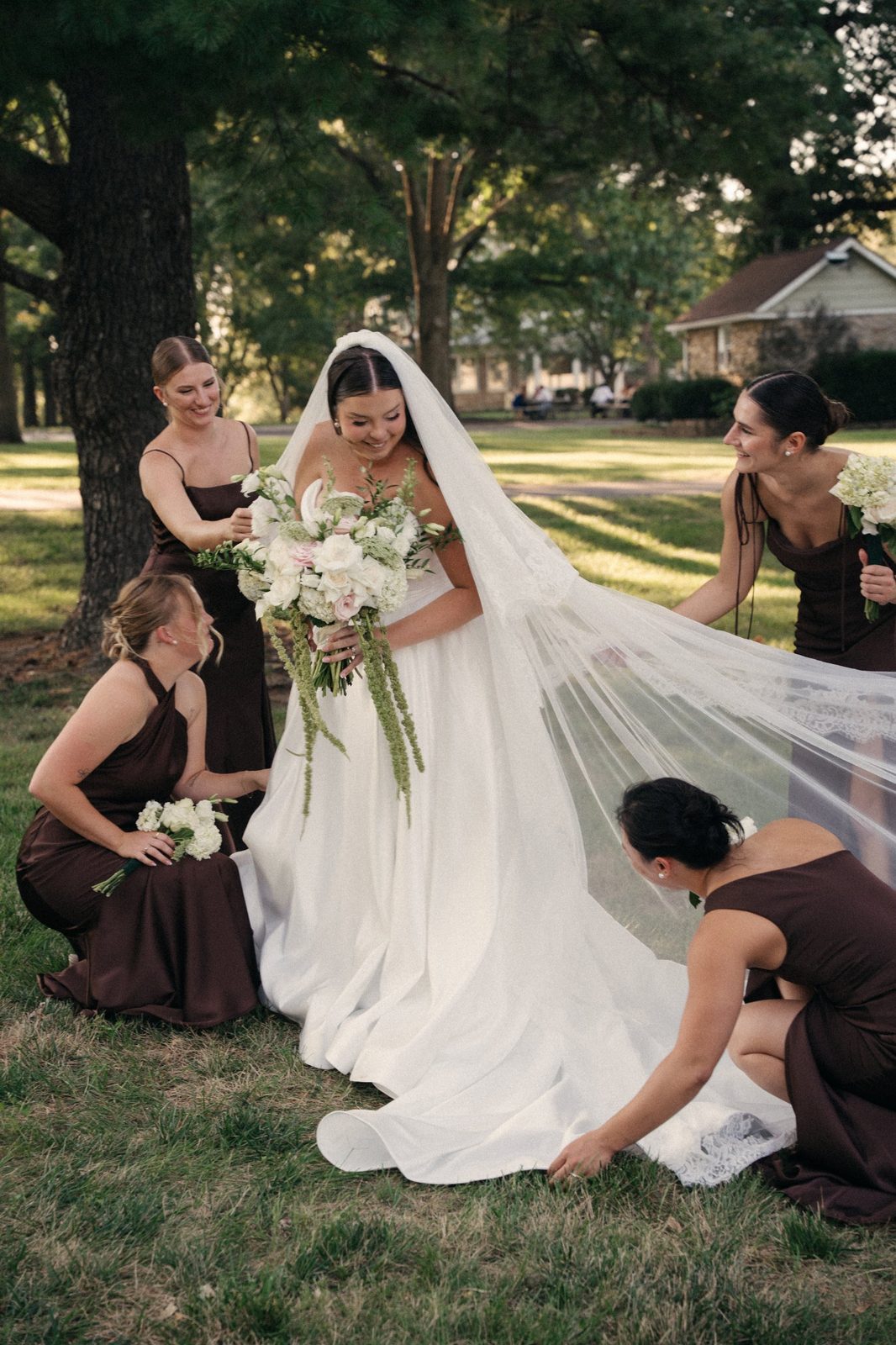Trinity smiling down at her bouquet while her bridesmaids in brown dresses fluff her long veil in the grass