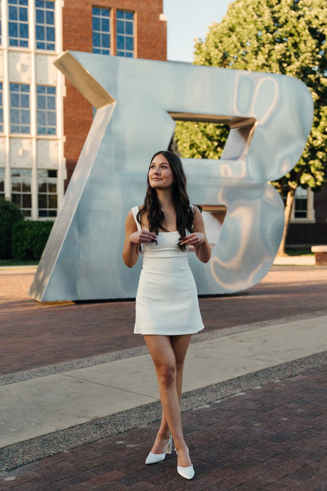 Charlotte in a white dress looking off to the side in front of the big silver B statue at Boise State