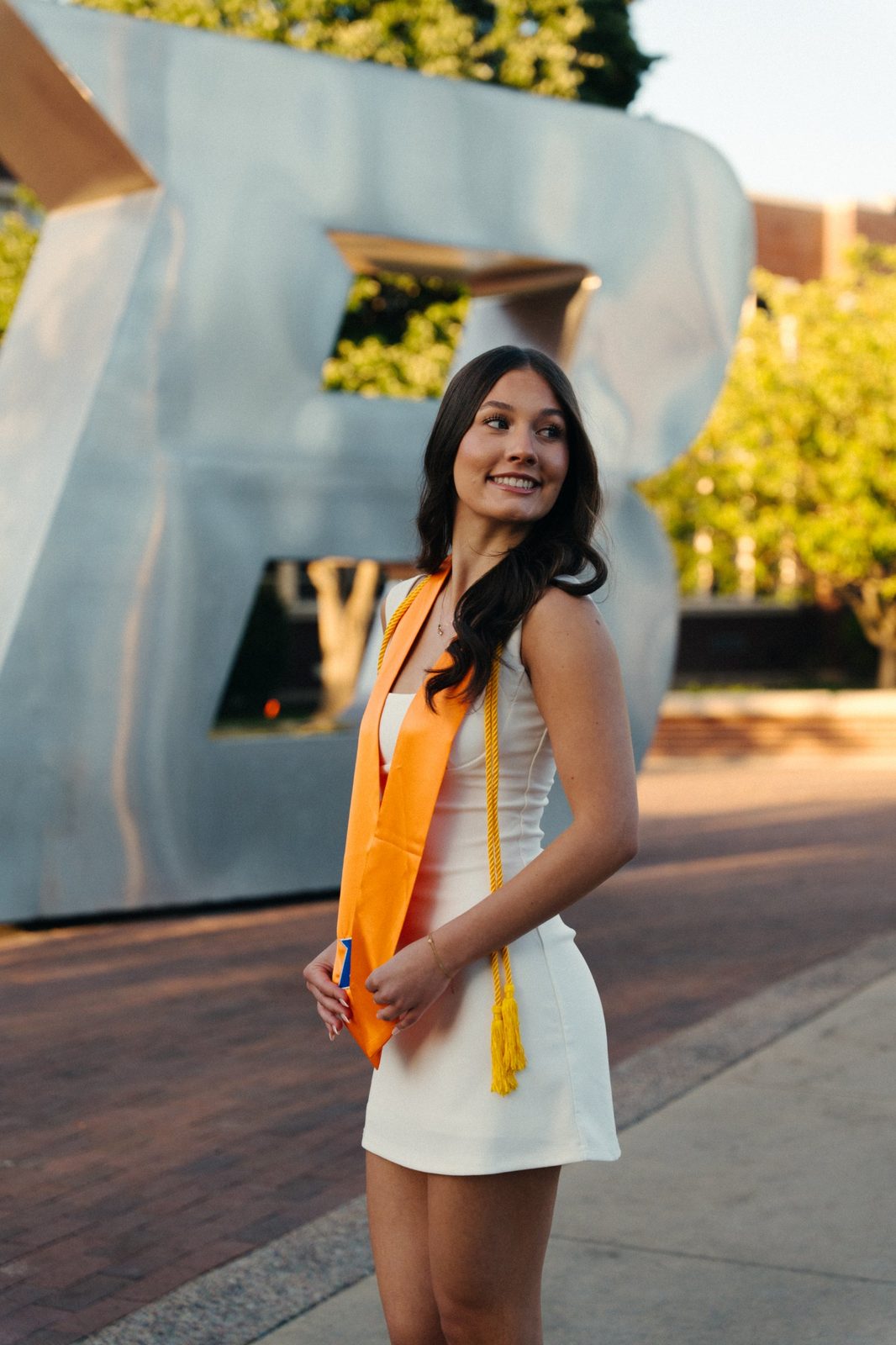 Charlotte in her stole and gold cords smiling over her shoulder in front of the silver B statue at Boise State