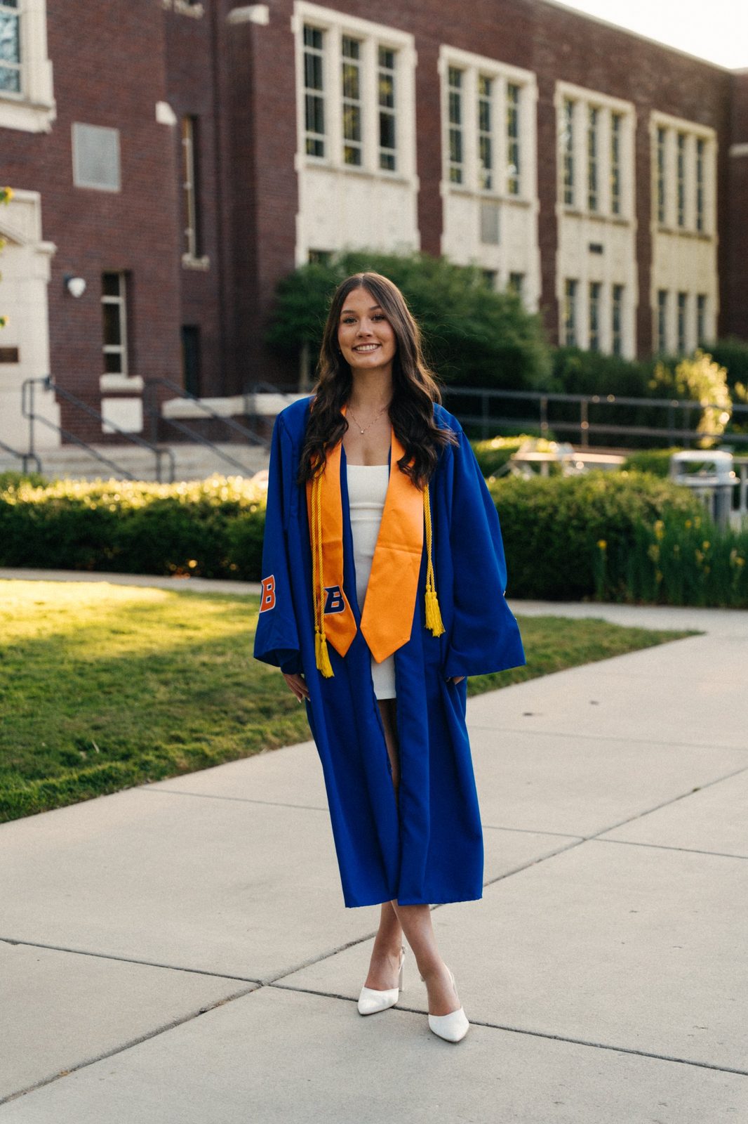 Charlotte in a blue Boise State graduation gown and orange stole standing on a campus sidewalk at golden hour