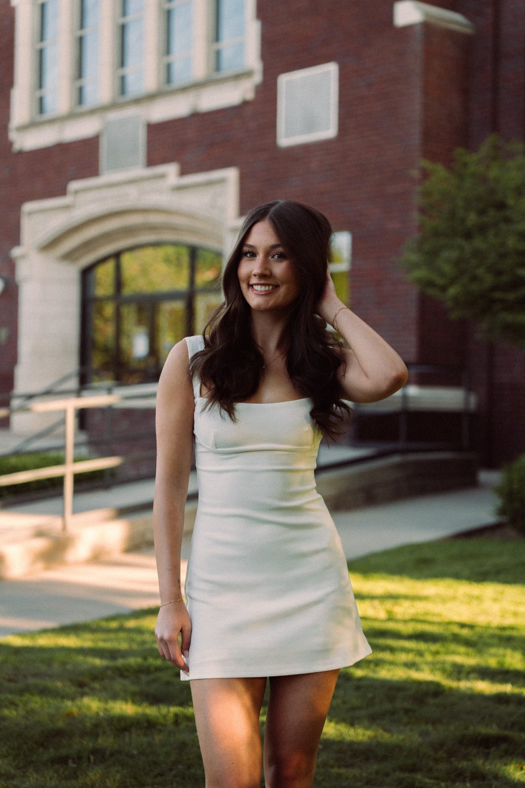 Charlotte smiling with her hand in her hair in a white dress in front of a brick building on the Boise State campus