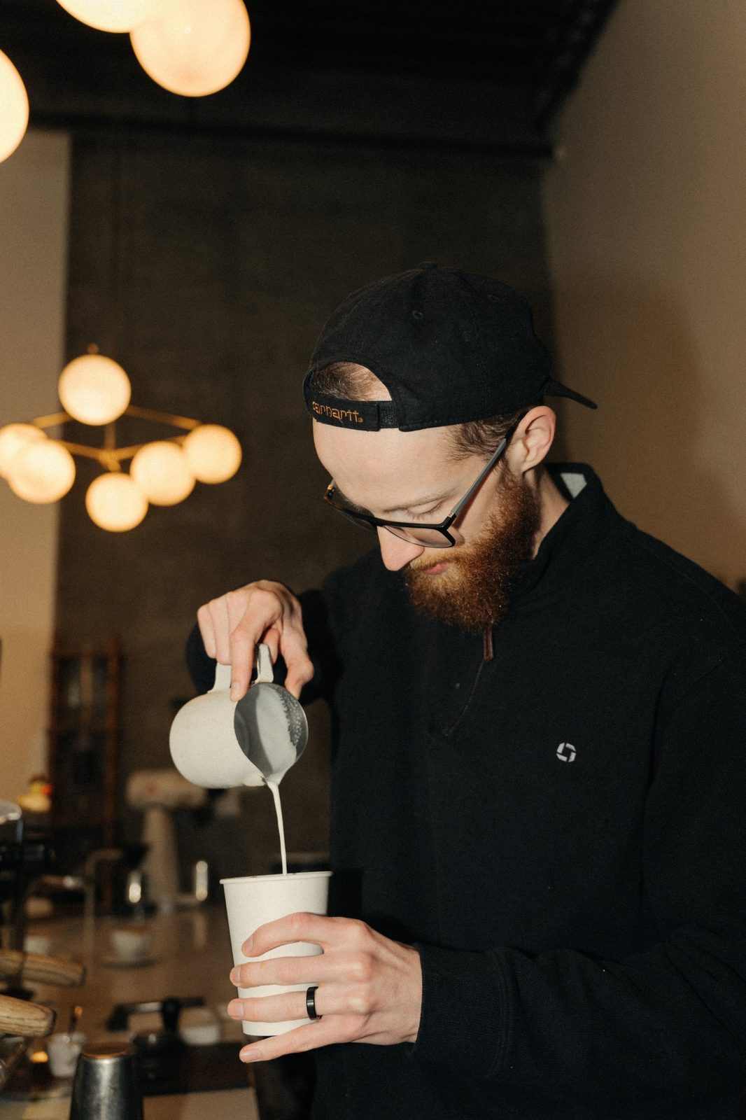 Barista in a backwards cap and glasses pouring steamed milk into a paper cup at a coffee bar with warm globe lights behind him