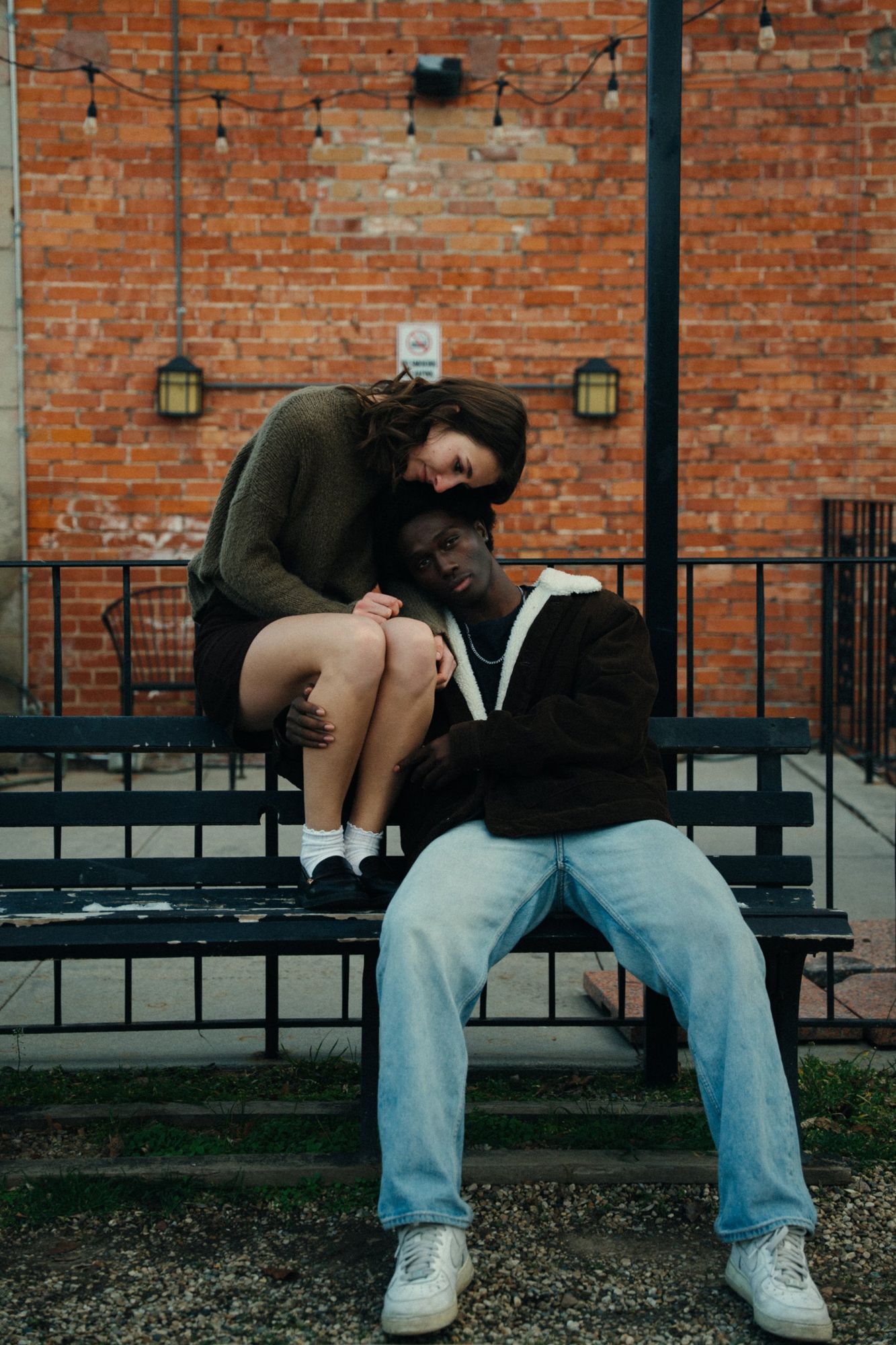 A couple sitting on a black bench in front of a red brick wall, she rests her head on his while he looks at the camera