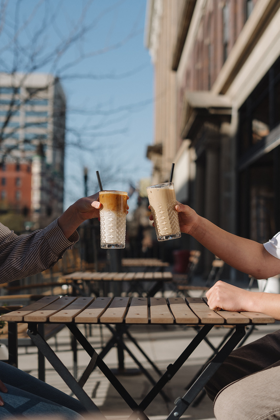 Two hands clinking iced lattes over a wooden patio table on a sunny downtown Boise street
