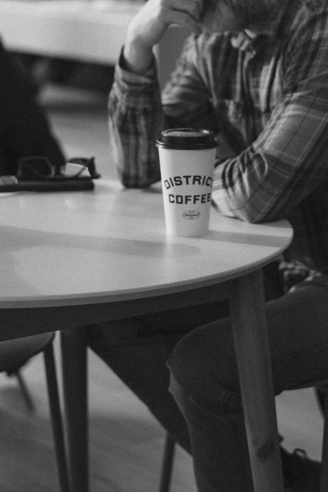 Black and white photo of a District Coffee cup on a round white table with a person in a plaid shirt beside it