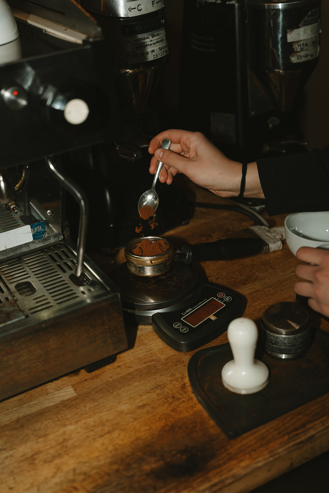 Barista spooning freshly ground coffee into a portafilter on a scale beside the espresso machine and grinders