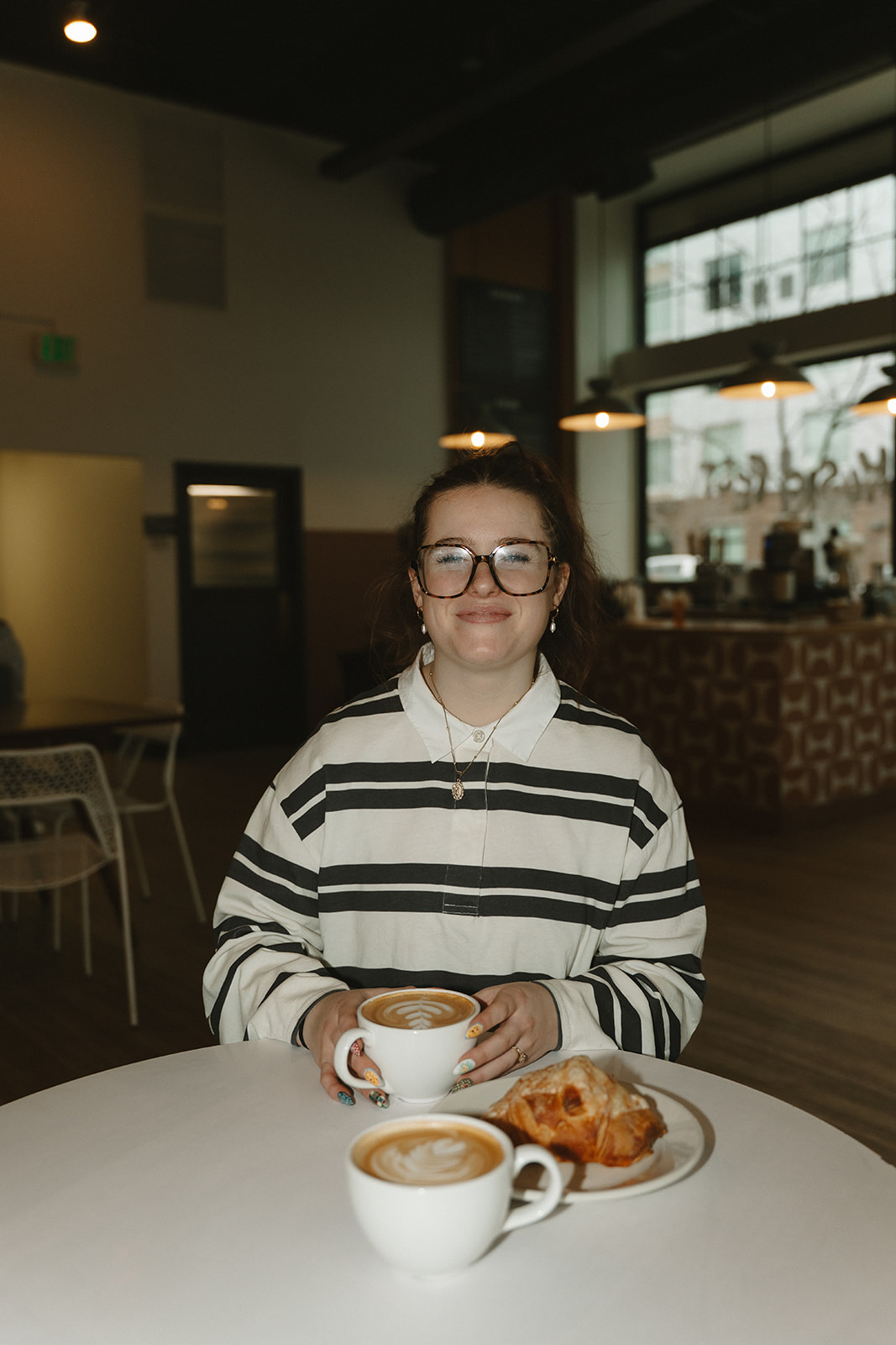 Woman in a striped rugby shirt and tortoiseshell glasses smiling at a table with two lattes and an almond croissant