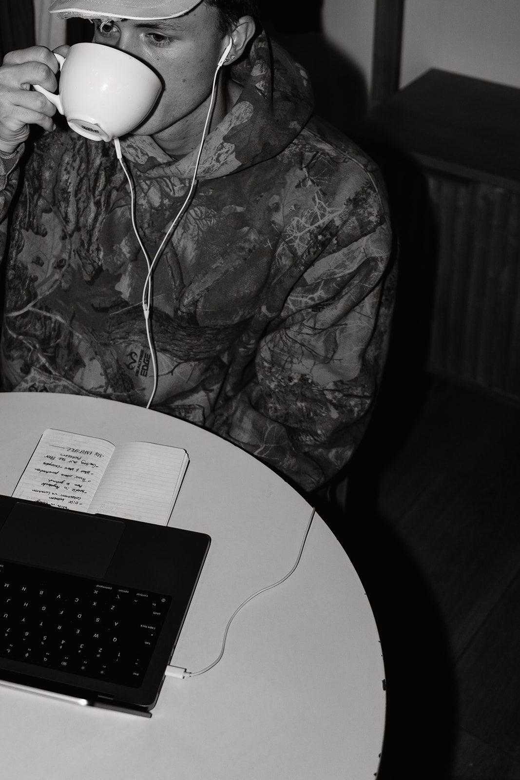 Black and white photo of a man in a camouflage hoodie sipping from a large cup beside a notebook and laptop
