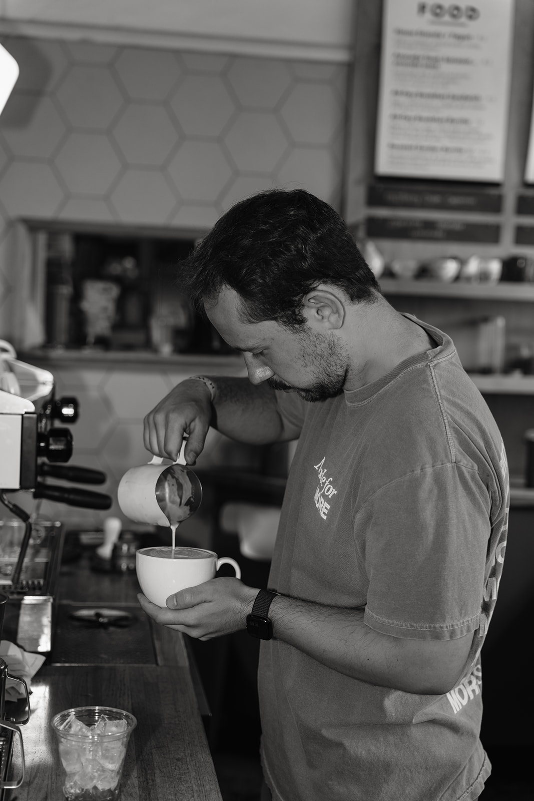 Black and white photo of a barista pouring a latte, bent over the cup in front of the hexagon tile wall