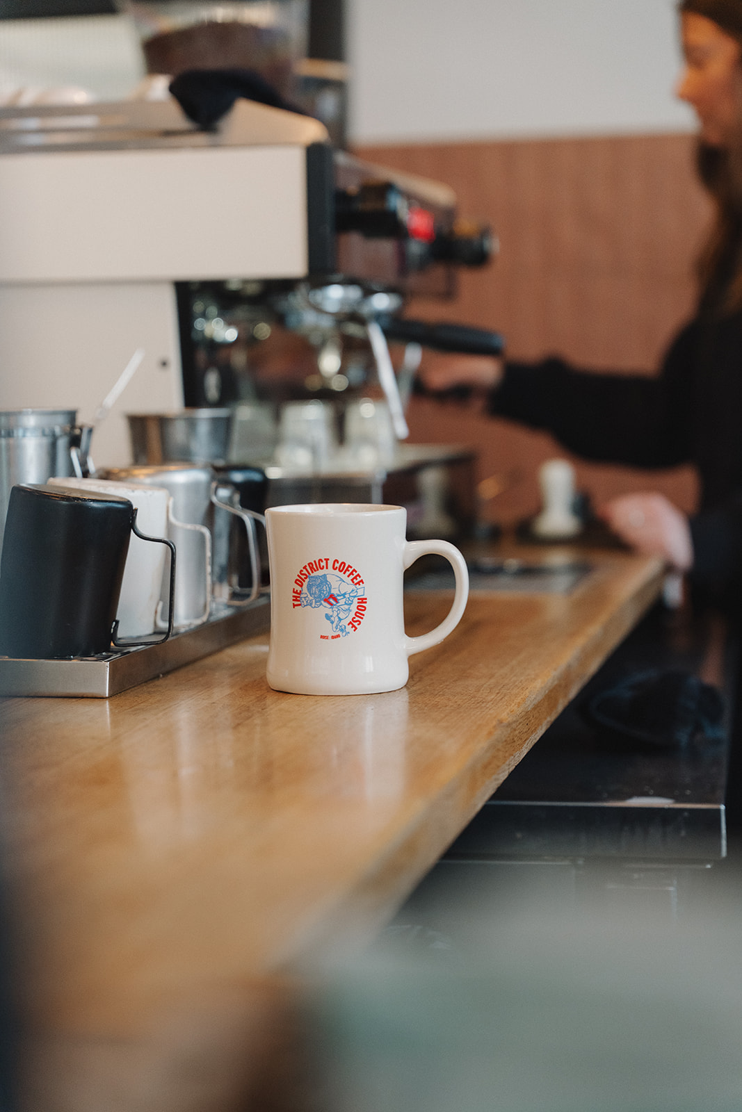 White District Coffee House mug with a red and blue Boise, Idaho logo sitting on a wooden bar next to steaming pitchers and an espresso machine