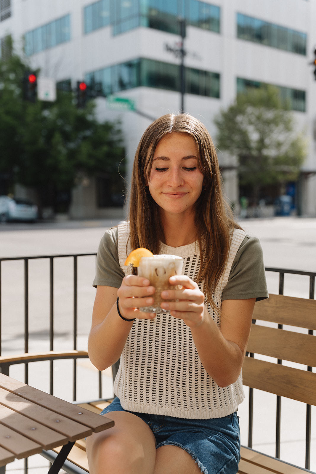 Woman smiling down at an iced coffee with an orange slice while sitting on a patio bench outside the District Coffee House