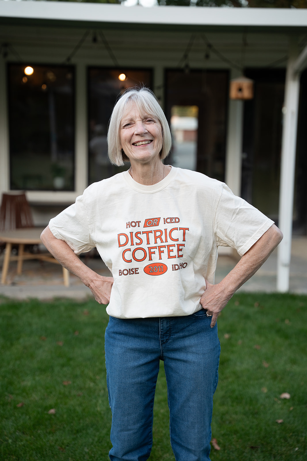 Older woman with a bright smile and her hands on her hips in a cream District Coffee Hot or Iced t-shirt, standing on a lawn
