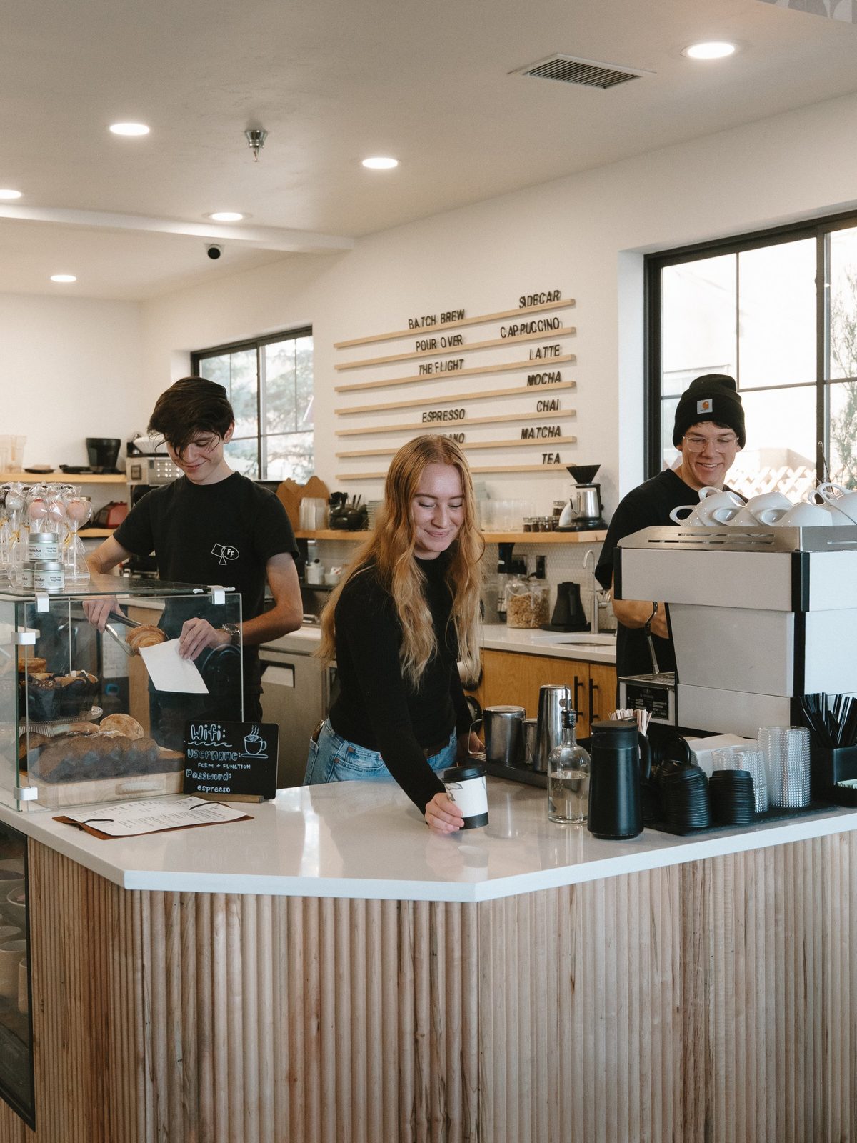 Three baristas smiling behind the counter at Form & Function, one picking up a croissant, one setting down a cup, one at the espresso machine