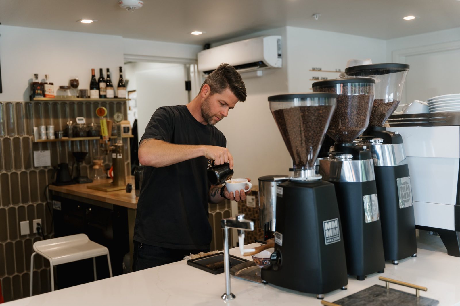 Barista pouring steamed milk into a cup beside three grinders full of coffee beans
