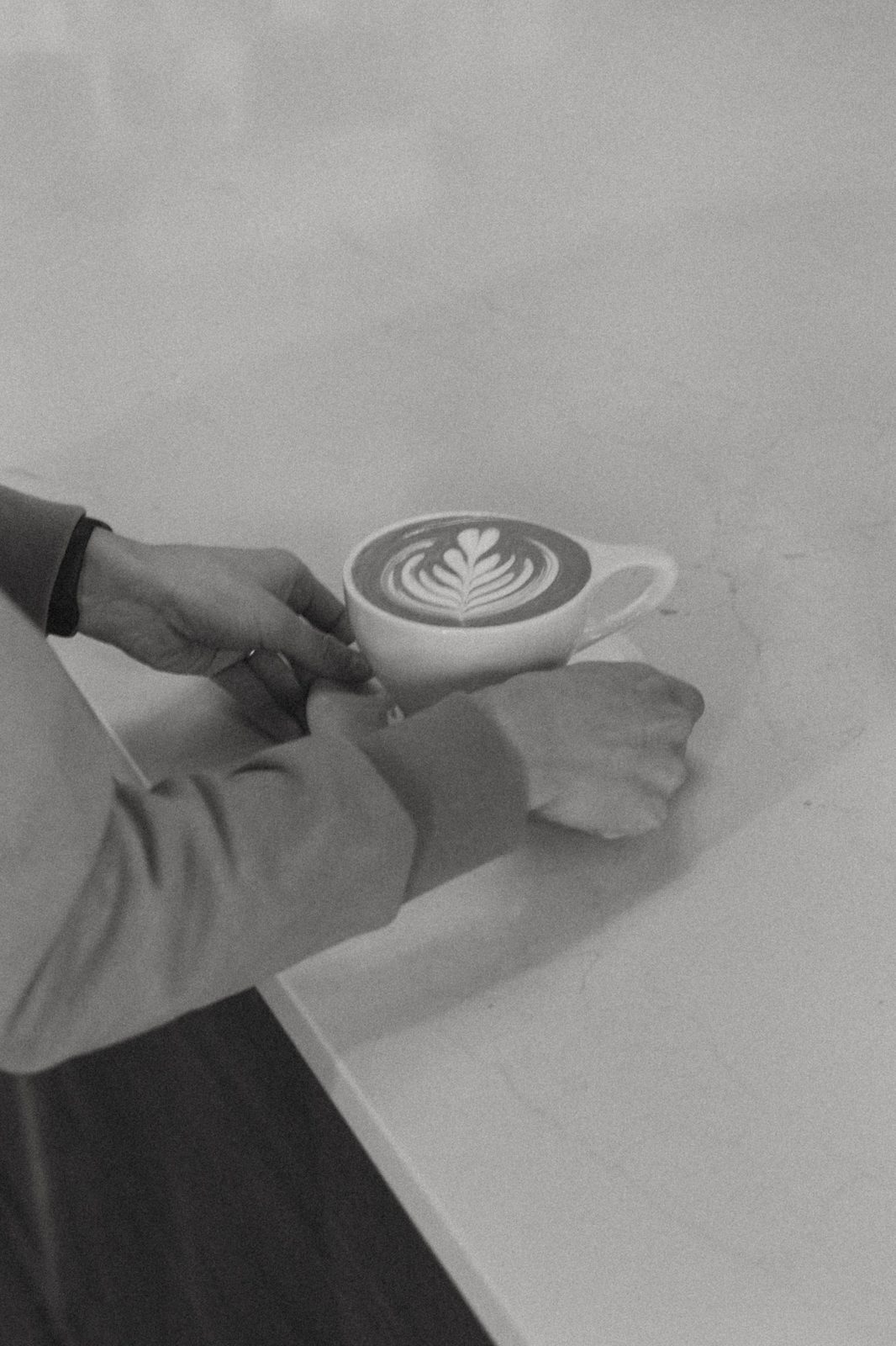 Black and white close-up of hands holding a latte with a rosetta pattern on a counter