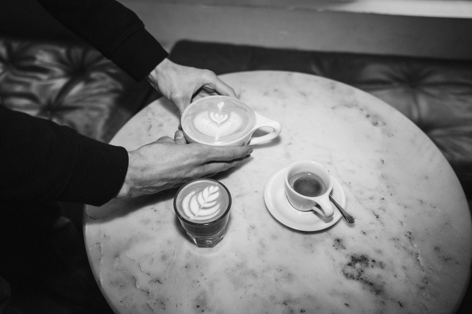 Black and white overhead of hands around a latte on a round marble table, with a cortado and an espresso beside it